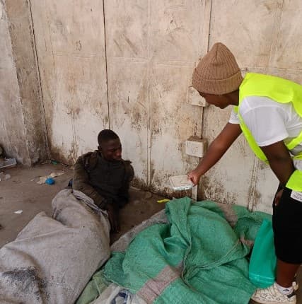 Volunteer in a neon vest hands a meal to a man sitting on the ground.