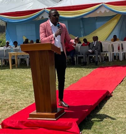 Man in a pink blazer speaking at a wooden podium on a red carpet outdoors.