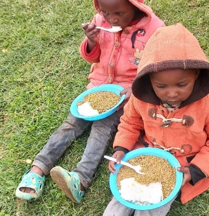 Two young children in hooded coats sit on green grass eating rice and lentils.