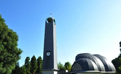 Tall clock tower with a cross beside modern domed architecture under a clear blue sky.